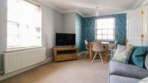 A room with a TV on a wooden stand, a dining table with chairs, a sofa, and a window with blinds.