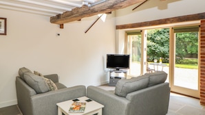 A living room with a grey sofa set, a small white table with a magazine, a flat-screen TV on a stand, and a large wooden beam ceiling. - The Cart Shed (Dereham)