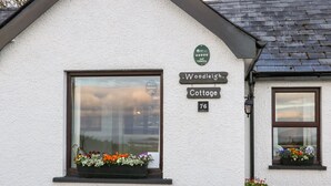 A white cottage with a window displaying a flower box. A sign above the window reads "Woodleigh Cottage 76". - Woodleigh Cottage (Ballycastle)