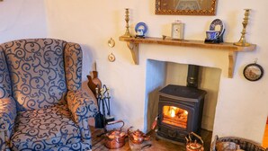 A cozy living room with a patterned armchair, a fireplace, a clock, and a shelf with decorative items. - Woodleigh Cottage (Ballycastle)