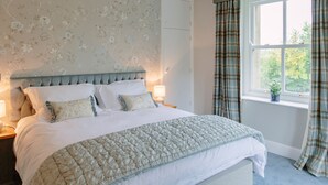 Une chambre à coucher avec une tête de lit capitonnée, un grand lit avec une literie blanche, un lustre, une fenêtre avec des rideaux à carreaux et un papier peint à motifs floraux.
