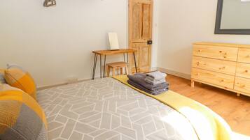 Un dormitorio con una cama, un tocador de madera, una mesita y una estantería con libros y un florero.