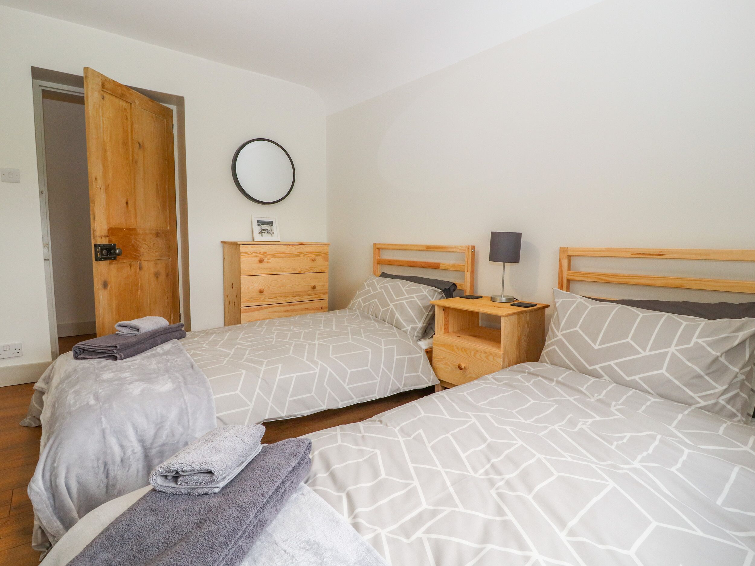 Un dormitorio con dos camas, cabeceros de madera, una mesita de noche con lámpara, un aparador y un espejo redondo en la pared.