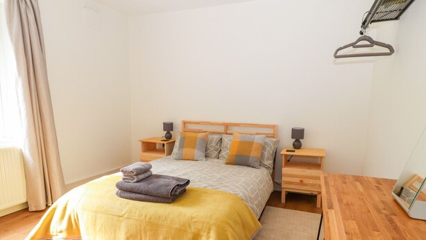 A neatly made bed with a yellow blanket, two bedside tables with lamps, a wooden nightstand, and a wooden chair. - Well House Farm Flat 1 (Chester)