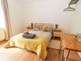 Una cama bien hecha con una colcha amarilla, dos mesitas de noche con lámparas, una mesita auxiliar de madera y una silla de madera.