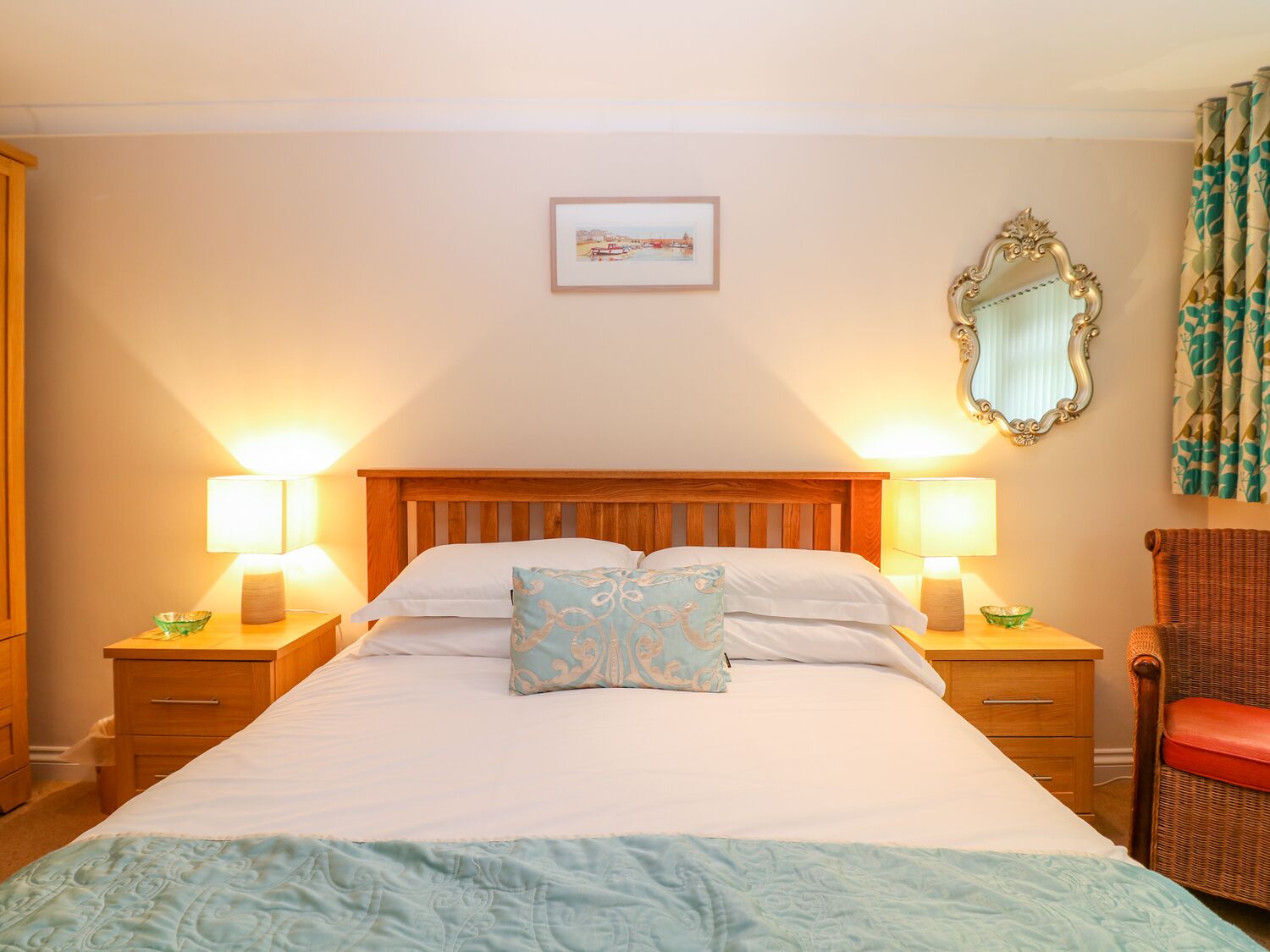 Une chambre d’hôtel bien aménagée, avec un grand lit, deux tables de chevet avec des lampes, un miroir et un fauteuil en osier.