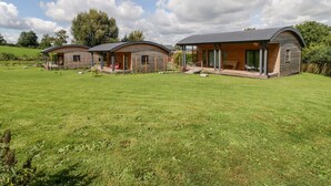 A row of three wooden cabins with curved roofs, each featuring a porch and a covered outdoor seating area. - Primrose (Shepton Mallet)