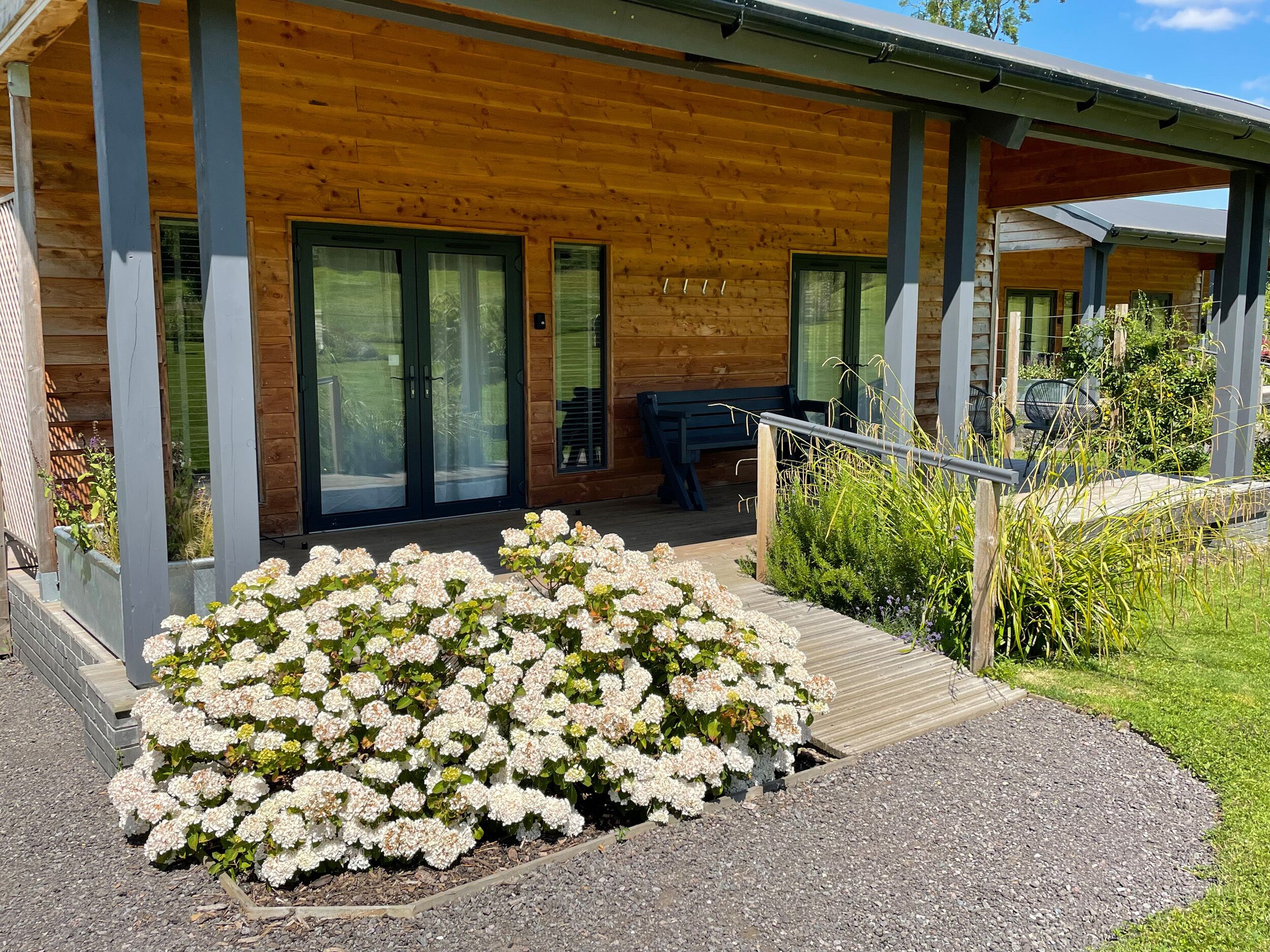 Eine Holzhütte mit überdachter Veranda, großen Schiebeg lassüren und einer kleinen Sitzgelegenheit im Freien.