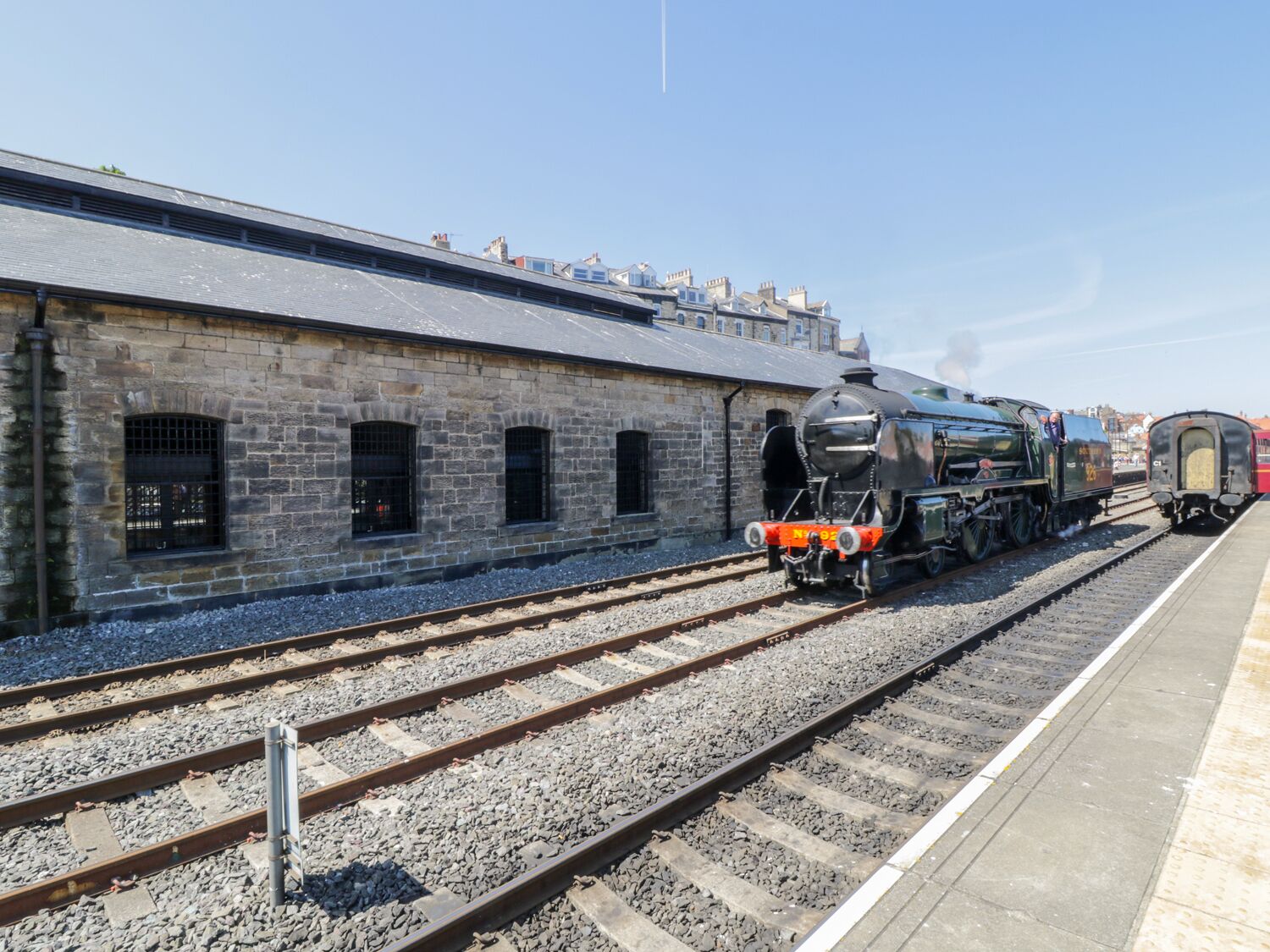Una estación de tren con un edificio de piedra, vías y una locomotora a vapor.