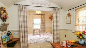 A cozy room with a bed, a table with a bottle and glasses, and a window with blinds. - Shepherds Hut - The Crook (Milford Haven)