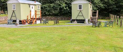 Two small, light green cottages with attached wooden structures, surrounded by a grassy area and trees.