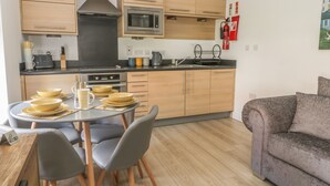 A modern kitchen with wooden cabinets, a stainless steel range hood, and a microwave. There is a dining table with chairs and a grey sofa in the living area. - The Krusty Krab (Filey)