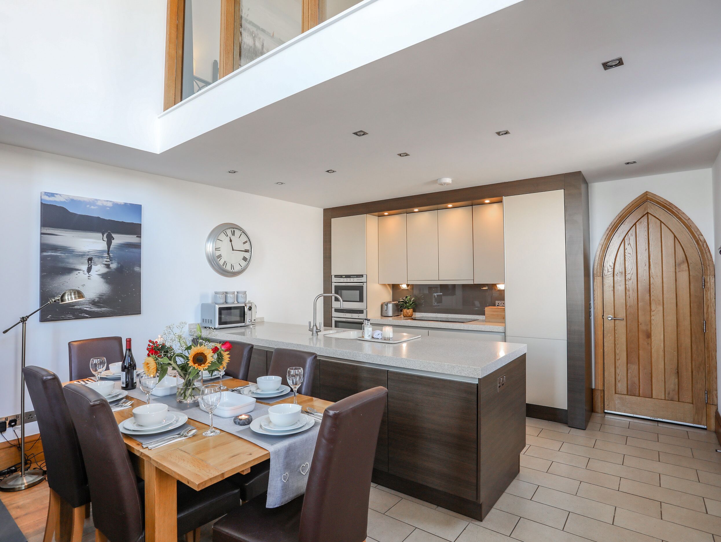 A modern kitchen with a central island, a dining area with a wooden table and chairs, a clock on the wall, and a large wooden door.
