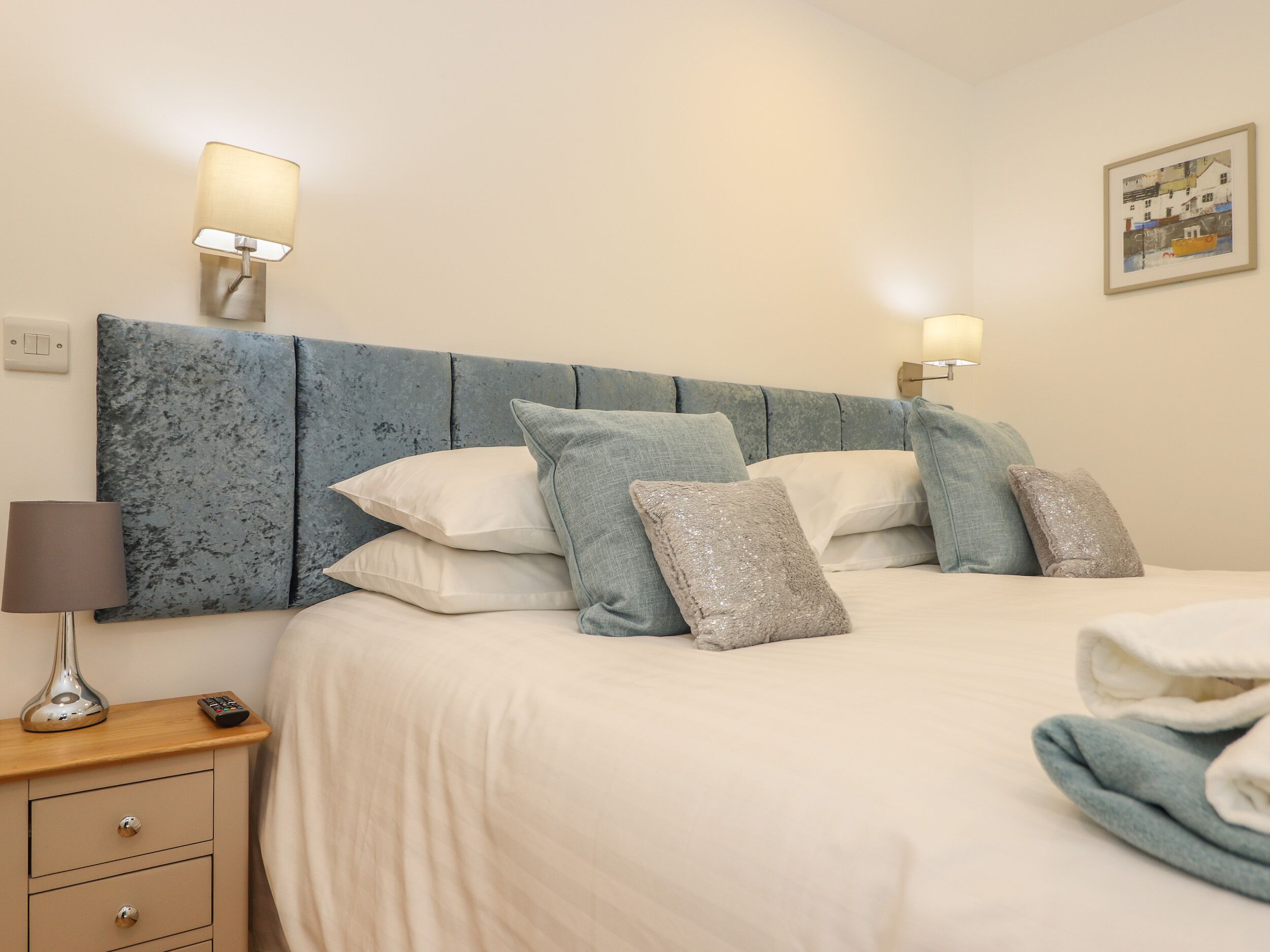 A neatly made bed with a blue headboard, multiple pillows, a bedside lamp, and a small wooden nightstand with a remote control.