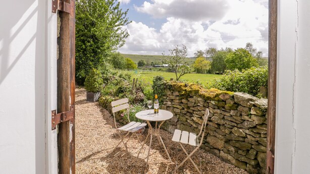 Une porte ouverte donnant sur un patio avec un mur en pierre, deux chaises blanches, une petite table ronde et une bouteille de vin.