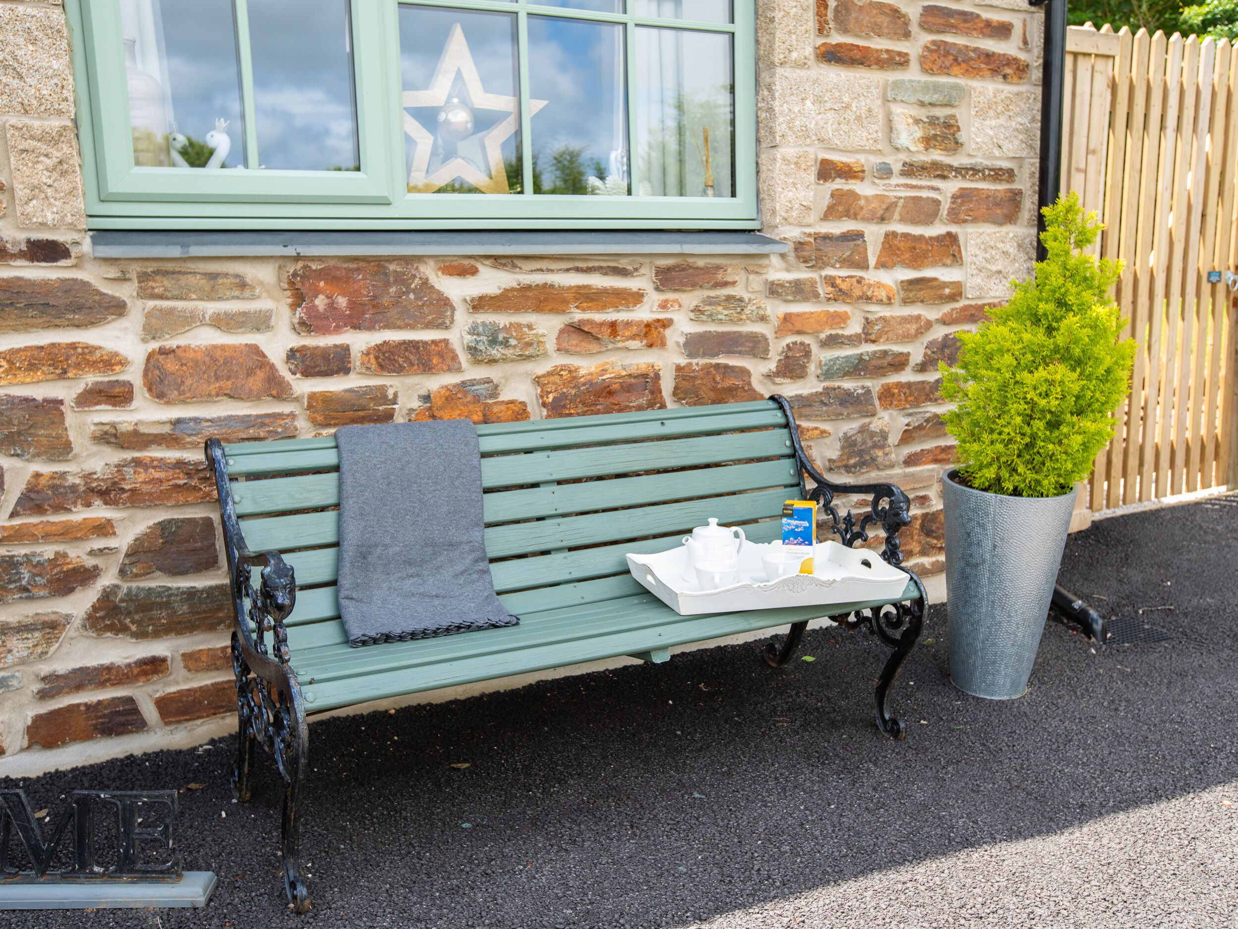 A green bench with a grey cloth, a white tray with a teapot and cups, and a potted plant beside a stone wall with a wooden fence.