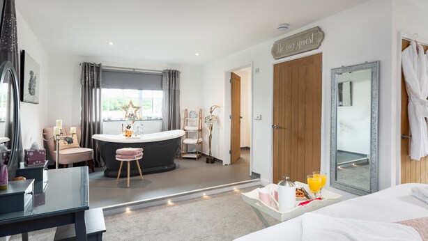 A hotel room with a bed, a desk, a vanity with a mirror, a bathtub, and a bathroom with a toilet and sink. - Be Our Guest Cottage (Truro)