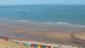 A beach with colorful beach huts and a ship in the distance. - Repton @ Engine Shed (Whitby)