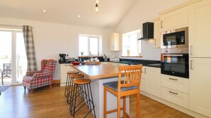 A modern kitchen with a central island, bar stools, and a dining area with a plaid chair. - Teal Cottage (Melton Mowbray)