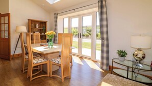 A dining area with a table, chairs, and a vase of flowers. There is a wooden cabinet and a glass table with a lamp. - Teal Cottage (Melton Mowbray)
