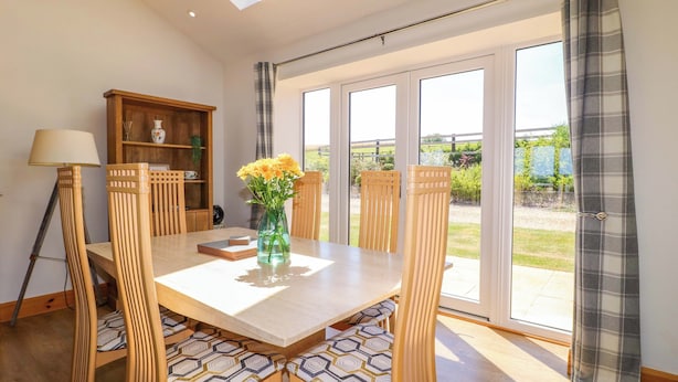 A dining area with a table, chairs, a vase with flowers, a lamp, and a view through large windows. - Teal Cottage (Melton Mowbray)