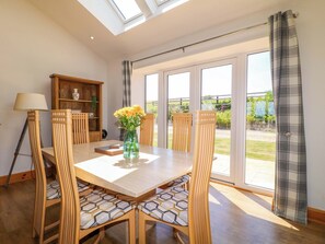 A dining area with a table, chairs, a vase with flowers, a lamp, and a view through large windows. - Teal Cottage (Melton Mowbray)