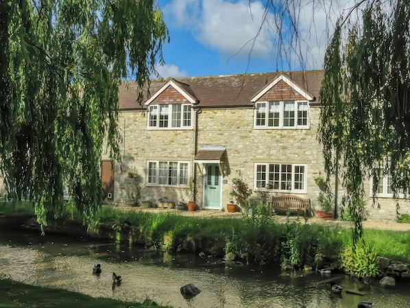 A stone house with a pond in front, surrounded by greenery and trees.