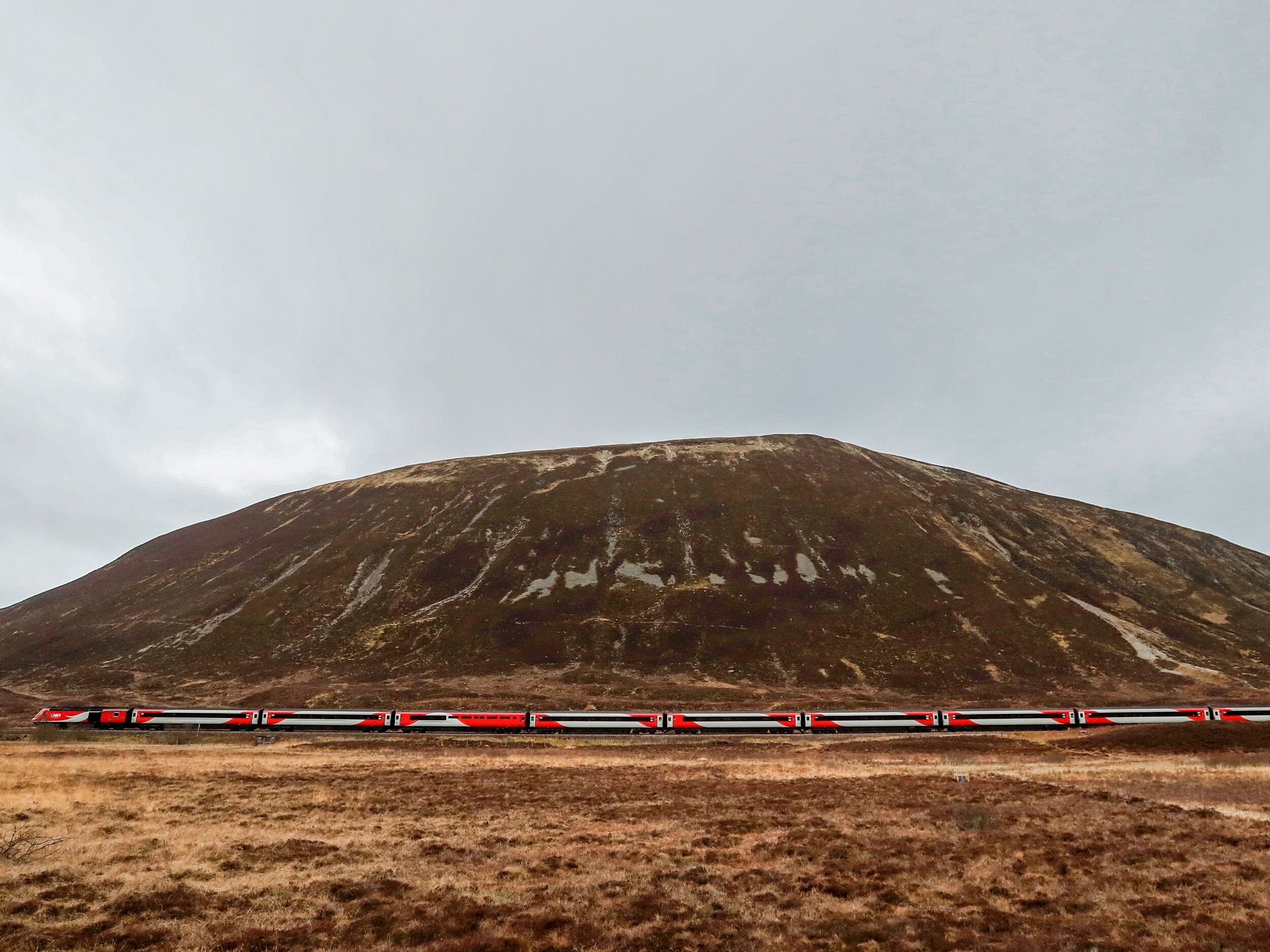 A train moving through a flat landscape with a large hill in the background.