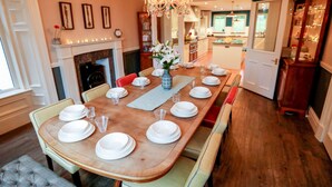 A dining room with a long wooden table set for six, flanked by chairs with cushioned seats. A chandelier hangs above the table, and a vase with flowers is placed in the center.