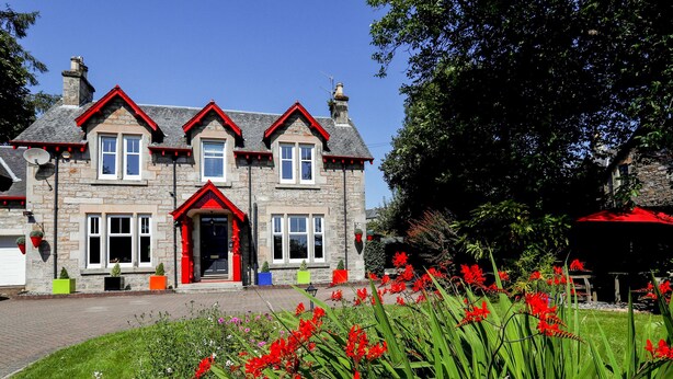 A stone building with a red entrance, multiple windows, and a well-maintained garden with red flowers.