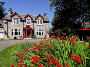 A stone building with a red entrance, multiple windows, and a well-maintained garden with red flowers. - Roseburn (Pitlochry)