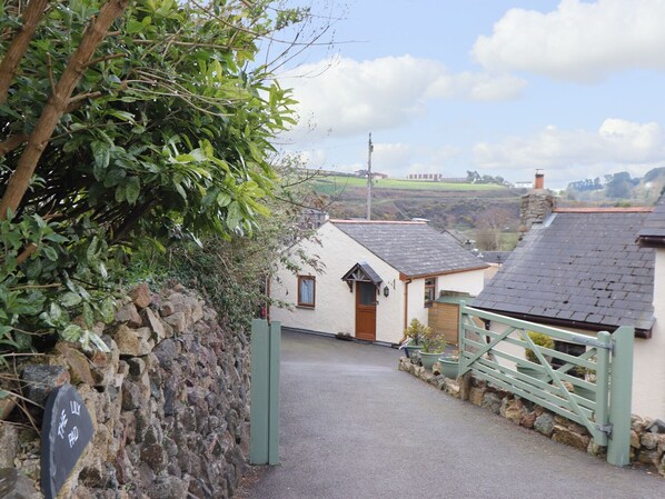 A narrow stone pathway leading to a white cottage with a green gate, surrounded by greenery and a rural landscape. - The Lily Pad (Camborne)