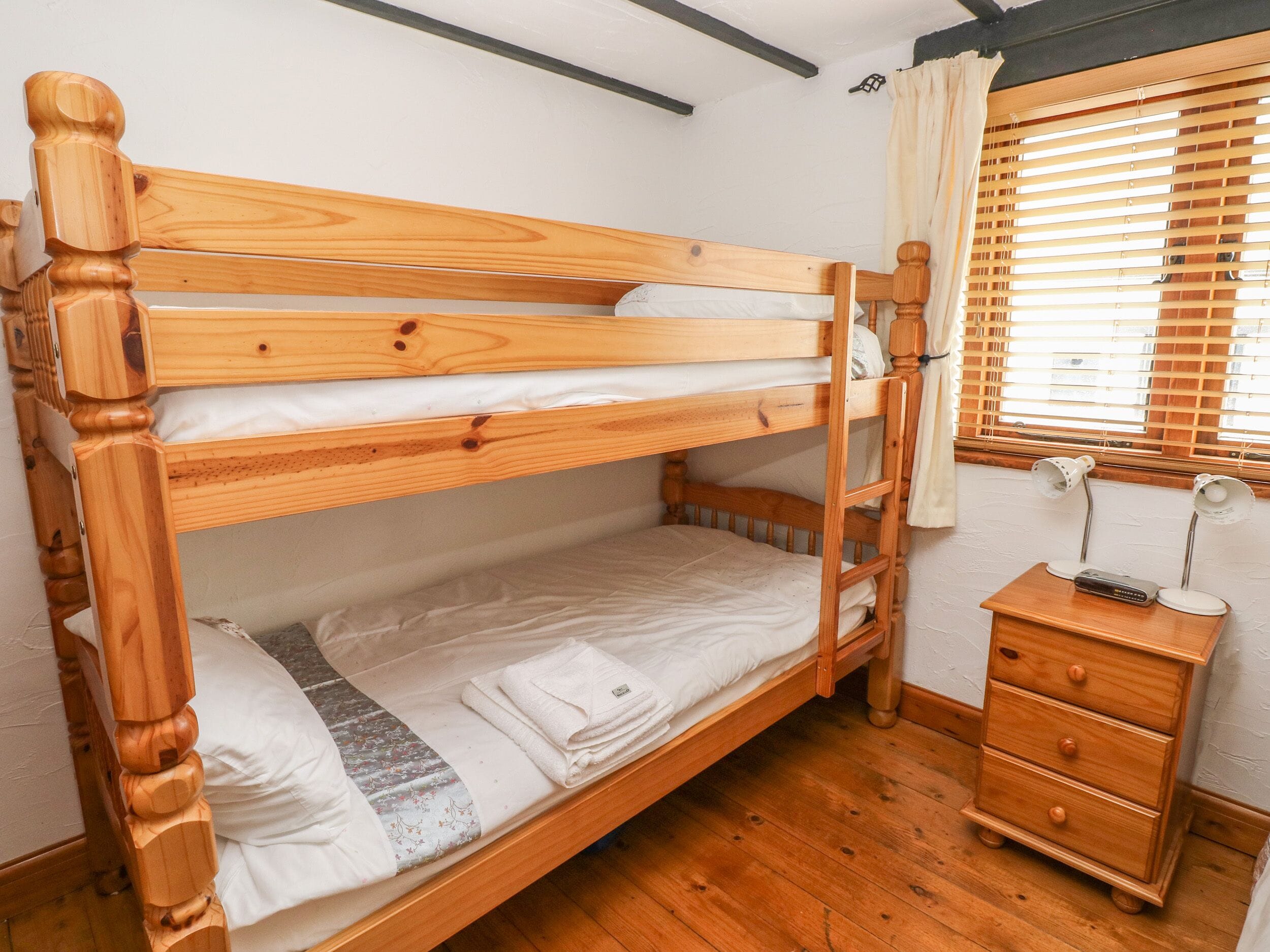 Een stapelbed met een trapje, een nachtkastje met een lamp en een raam met jaloezieën.