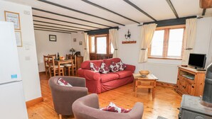 A cozy living room with a red sofa, two armchairs, a wooden table with a bowl, a television, and a wood-burning stove. - Cowslip Cottage (Milford Haven)