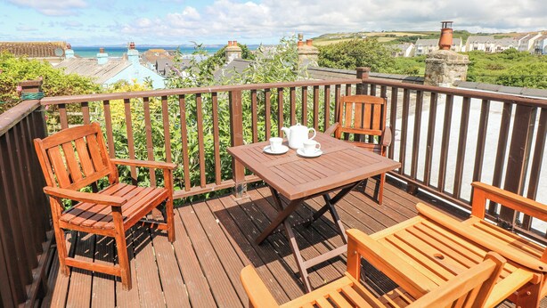 Une terrasse en bois avec une table et des chaises, offrant une vue sur un paysage côtier avec des maisons et la mer.