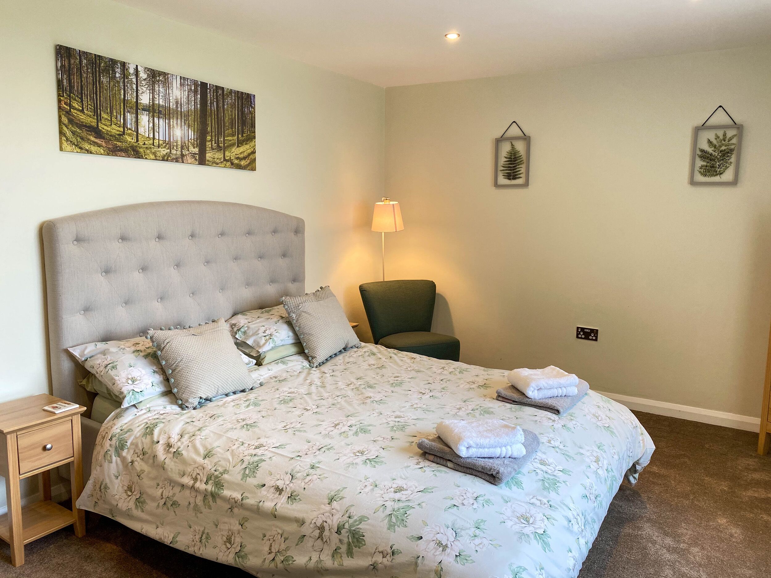 A neatly made bed with floral bedding, a tufted headboard, and two pillows. There is a small bedside table with a lamp, a chair, and a framed picture on the wall.