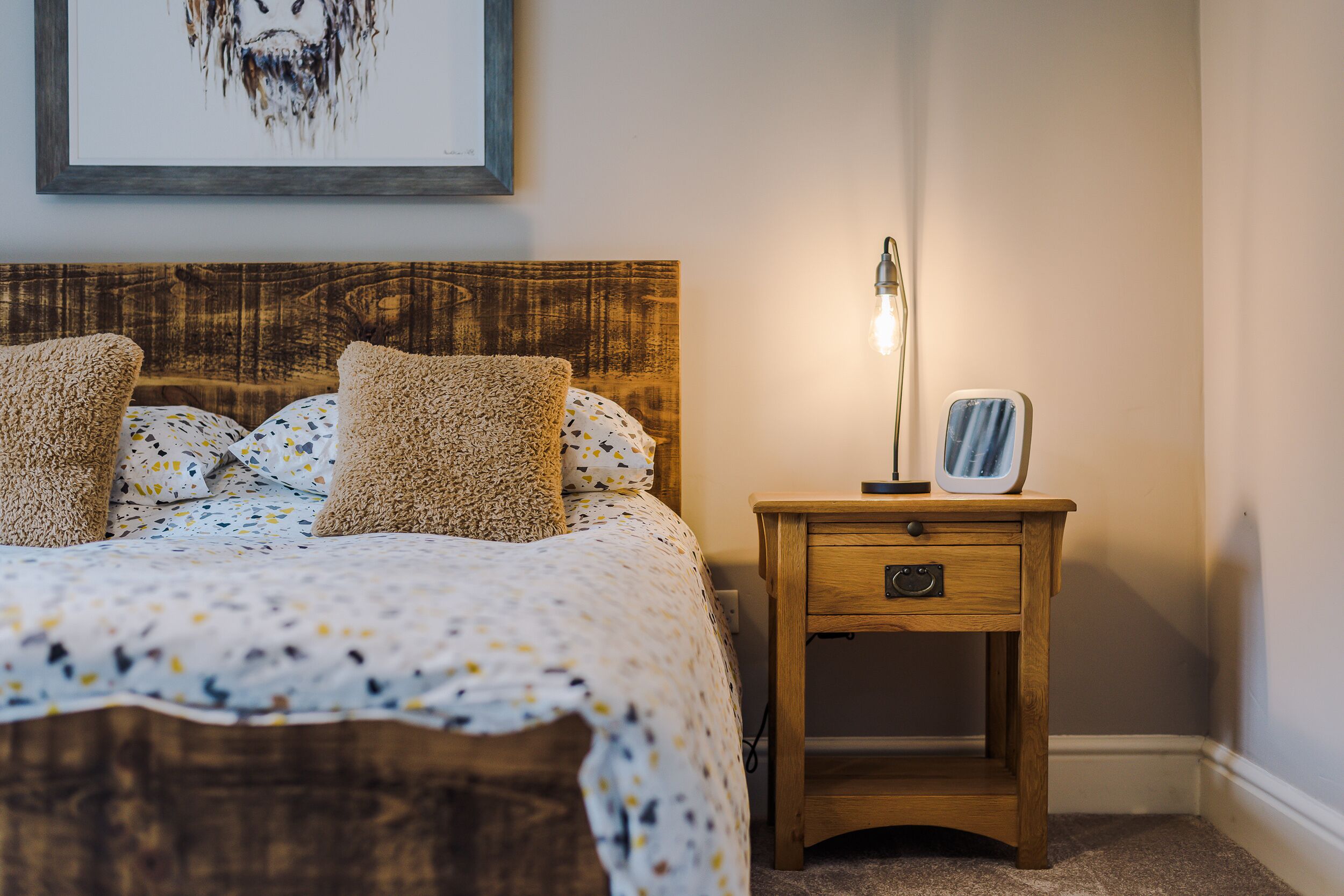 A neatly made bed with a patterned bedspread and two pillows, a wooden nightstand with a lamp and a framed picture, and a wall with a framed artwork.