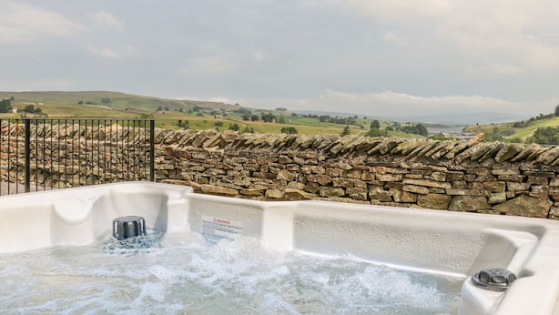 A hot tub with a stone wall and metal railing, overlooking a scenic landscape with hills and a body of water. - The Aspens (Barnard Castle)