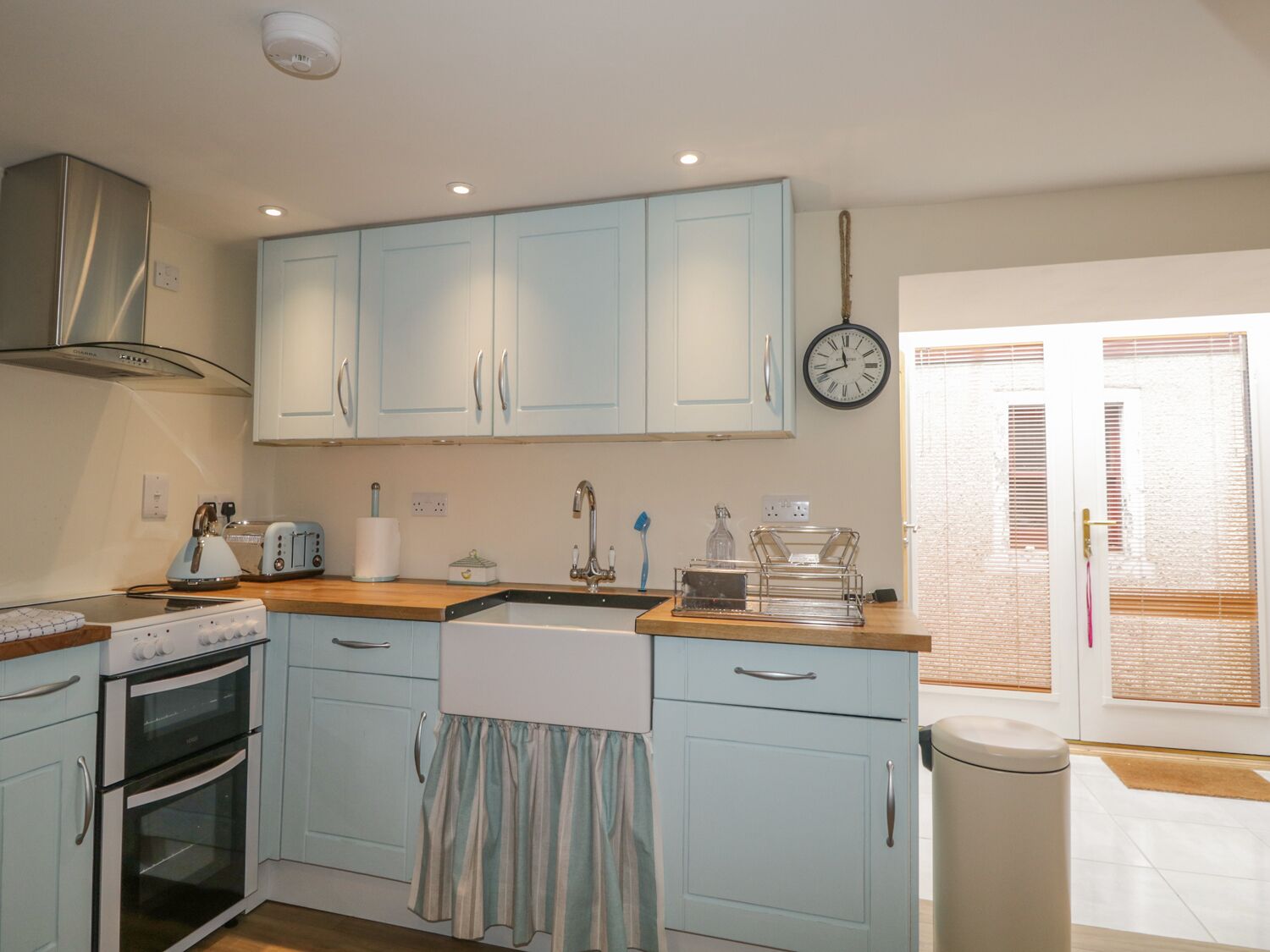 A kitchen with light blue cabinets, a stainless steel range hood, a double oven, a sink, and a clock on the wall.