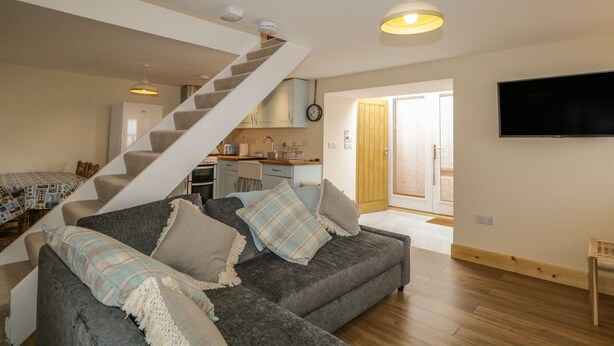 A modern living room with a grey sofa, a staircase, a kitchen, and a TV mounted on the wall. - 11 Village (Banff)