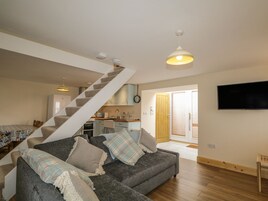 A modern living room with a grey sofa, a staircase, a kitchen, and a TV mounted on the wall.