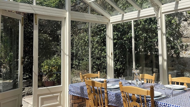 A conservatory with a table set for dining, surrounded by stone walls and greenery. - Sant Seiriol (Beaumaris)