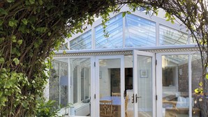 A conservatory with a glass roof, a wooden door, and a view of a dining area inside. - Sant Seiriol (Beaumaris)