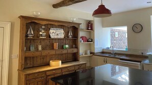 A kitchen with wooden cabinetry, a black countertop, a red pendant light, and a brick wall visible through a window. - Sant Seiriol (Beaumaris)