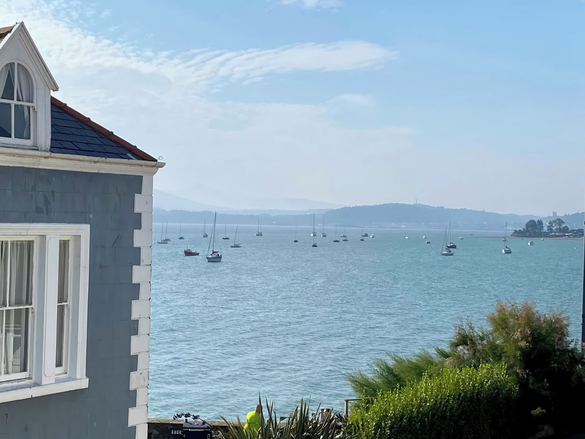 A coastal view with a building featuring a sloped roof and a bay with multiple sailboats.
