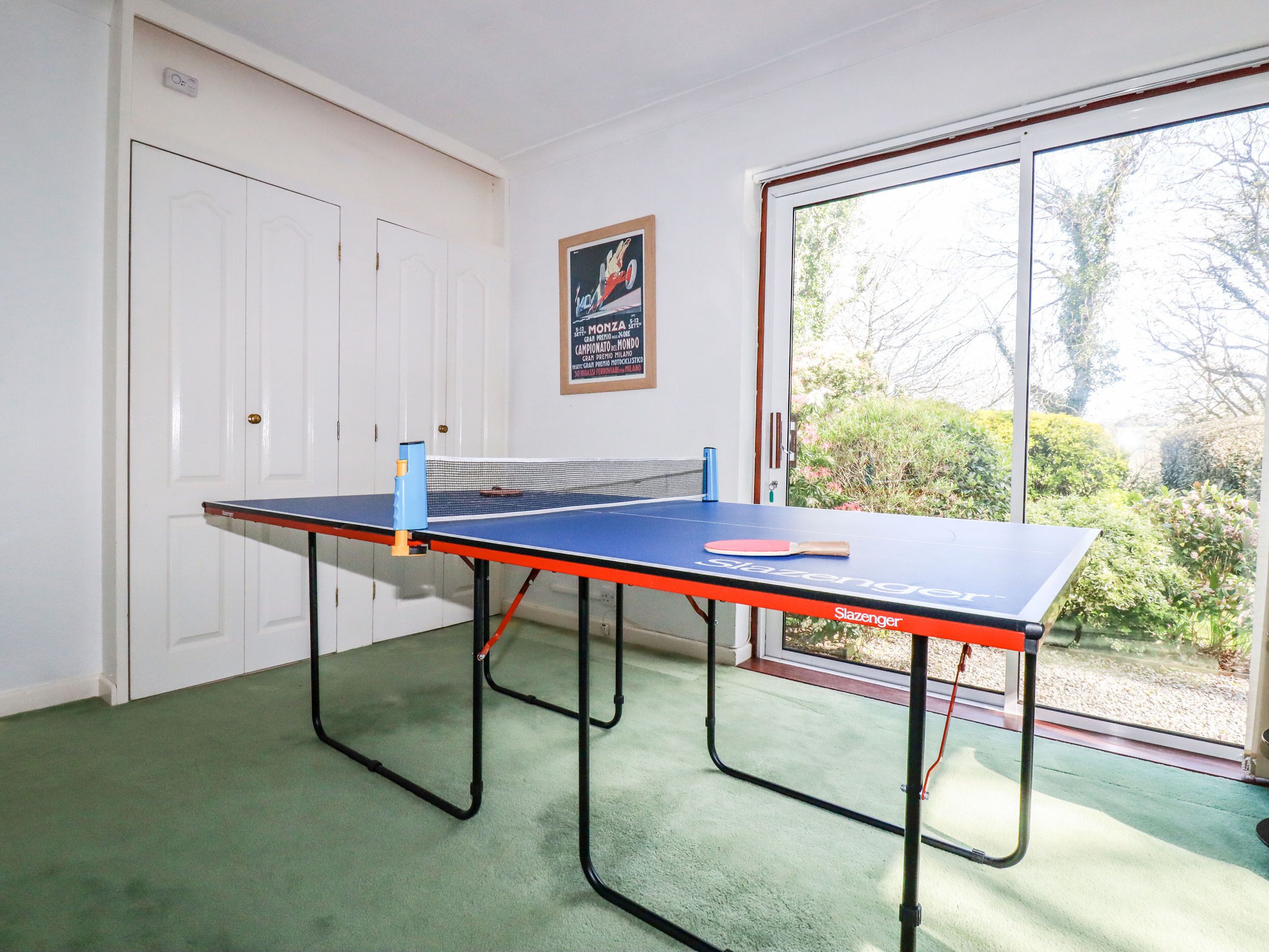 Ein Tischtennis-Tisch in einem Raum mit grünem Teppich, weißen Türen und einem Poster an der Wand.