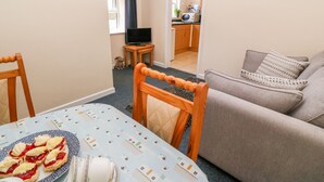 A cozy living room with a sofa, a dining table with food, and a kitchen in the background. - The Old Cows House (Helston)