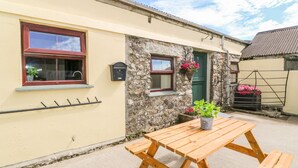 A yellow building with a stone wall, a wooden bench, and a small table with a potted plant. - The Old Cows House (Helston)