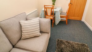 A cozy living room with a beige sofa, a patterned throw pillow, a small table with a floral centerpiece, and a wooden door. - The Old Cows House (Helston)