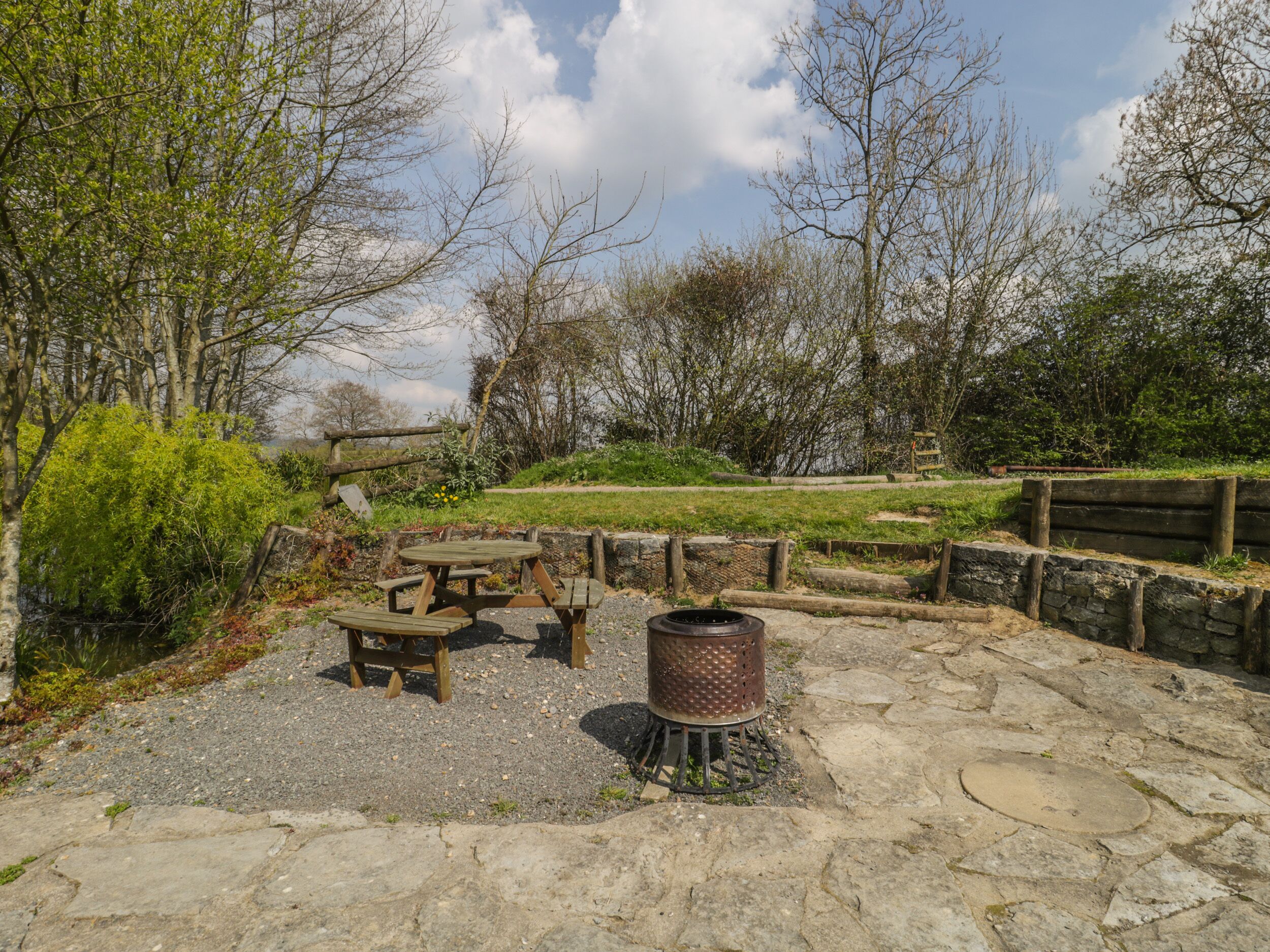 A rustic outdoor seating area with a wooden table and benches, surrounded by stone walls and greenery.